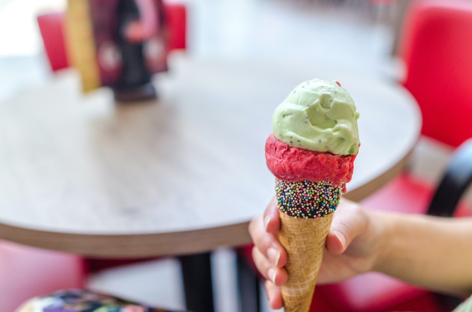 A two-scoop ice cream cone in a consumer’s hand at a table in an ice cream parlour